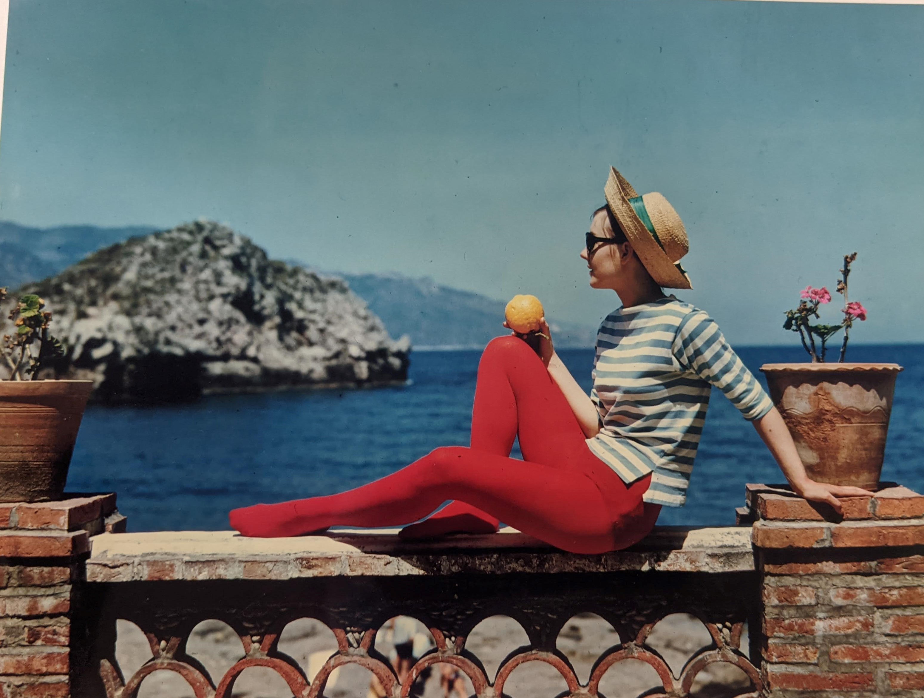 Woman wearing red pants, a blue and white striped shirt, and a straw hat sitting on a wall looking out at the water.